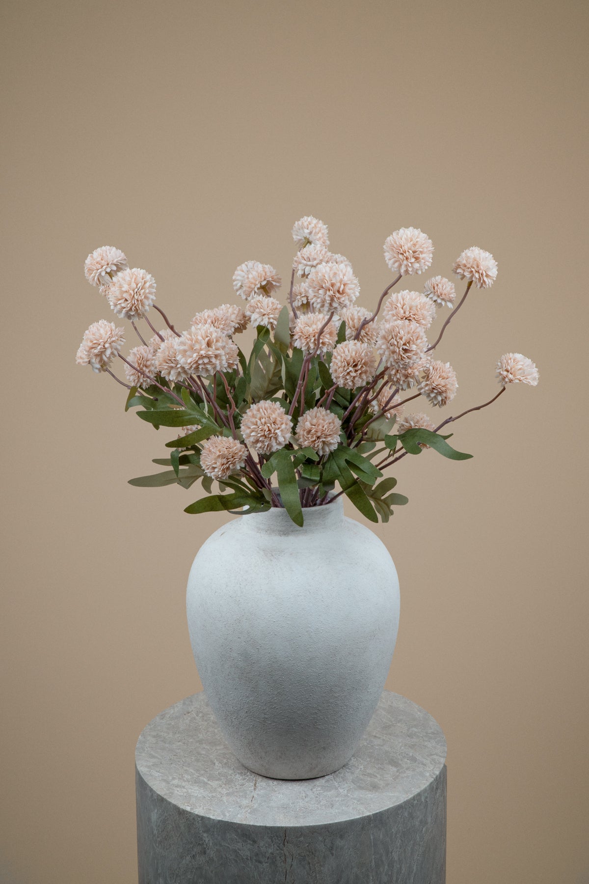 A dried floral arrangement with pale blush pink pom-pom flowers and green leaves in a matte white ceramic vase, displayed on a round gray pedestal against a warm beige background.