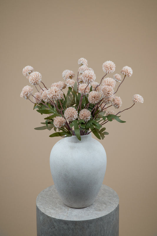 A dried floral arrangement with pale blush pink pom-pom flowers and green leaves in a matte white ceramic vase, displayed on a round gray pedestal against a warm beige background.