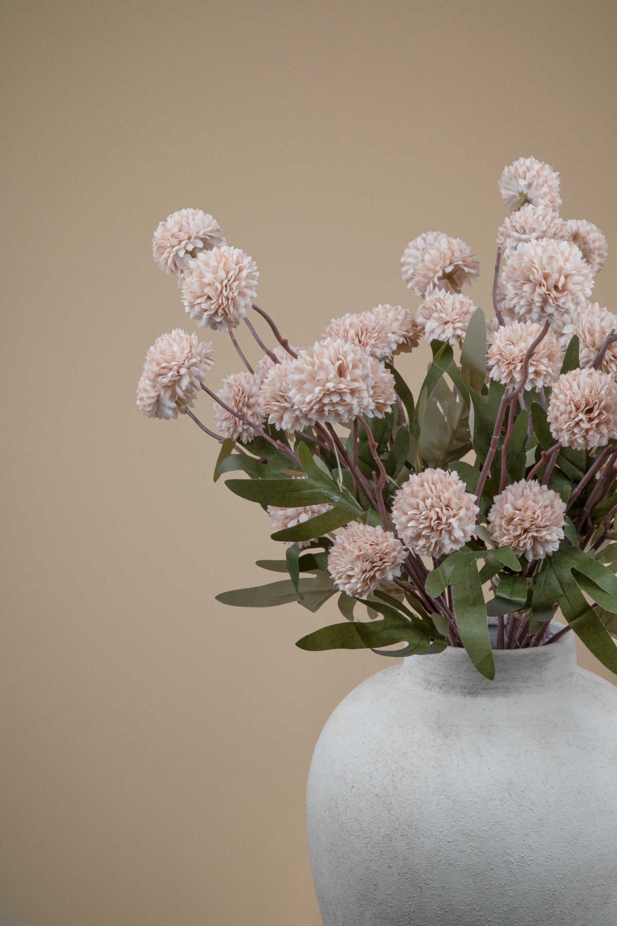A vase of dried decorative flowers with pale blush pink pom-pom blooms and green leaves, arranged in a textured off-white ceramic vase against a warm beige background.