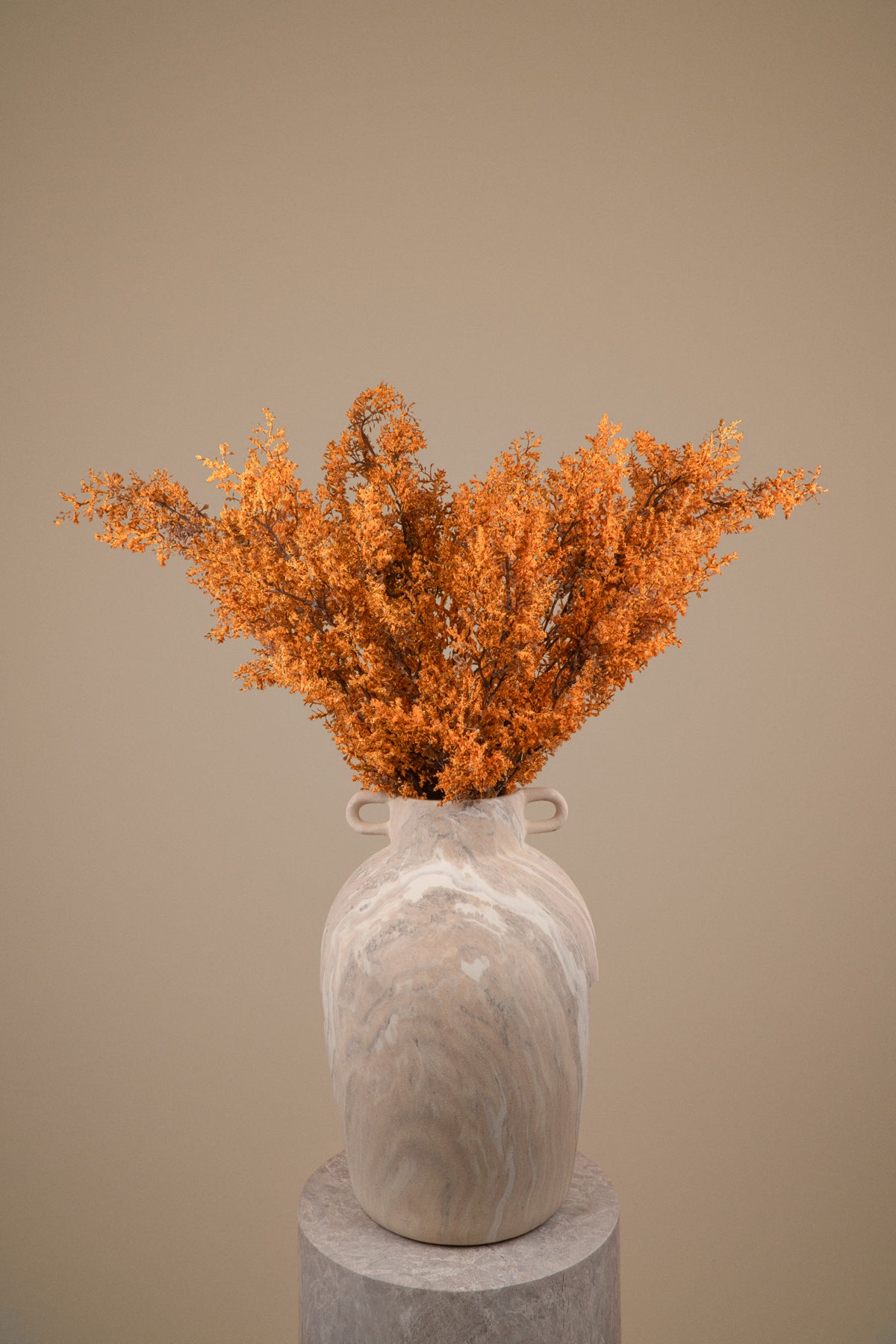 A decorative ceramic vase with a marbled light gray and white finish, holding a dense bouquet of dried orange foliage, displayed on a round pedestal against a solid beige background.
