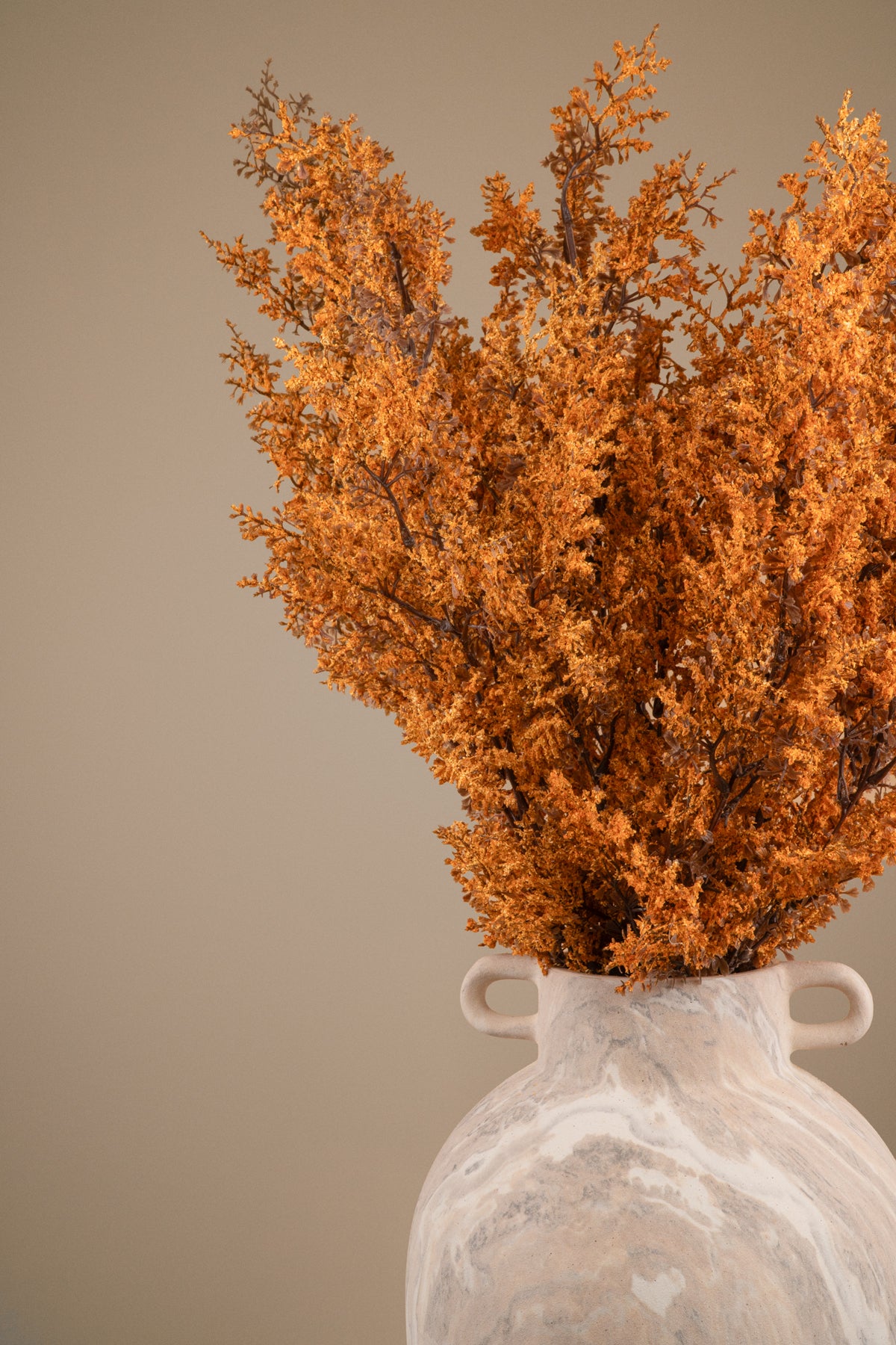 Dried orange foliage arranged in a white ceramic vase with two side handles, set against a plain warm beige background.