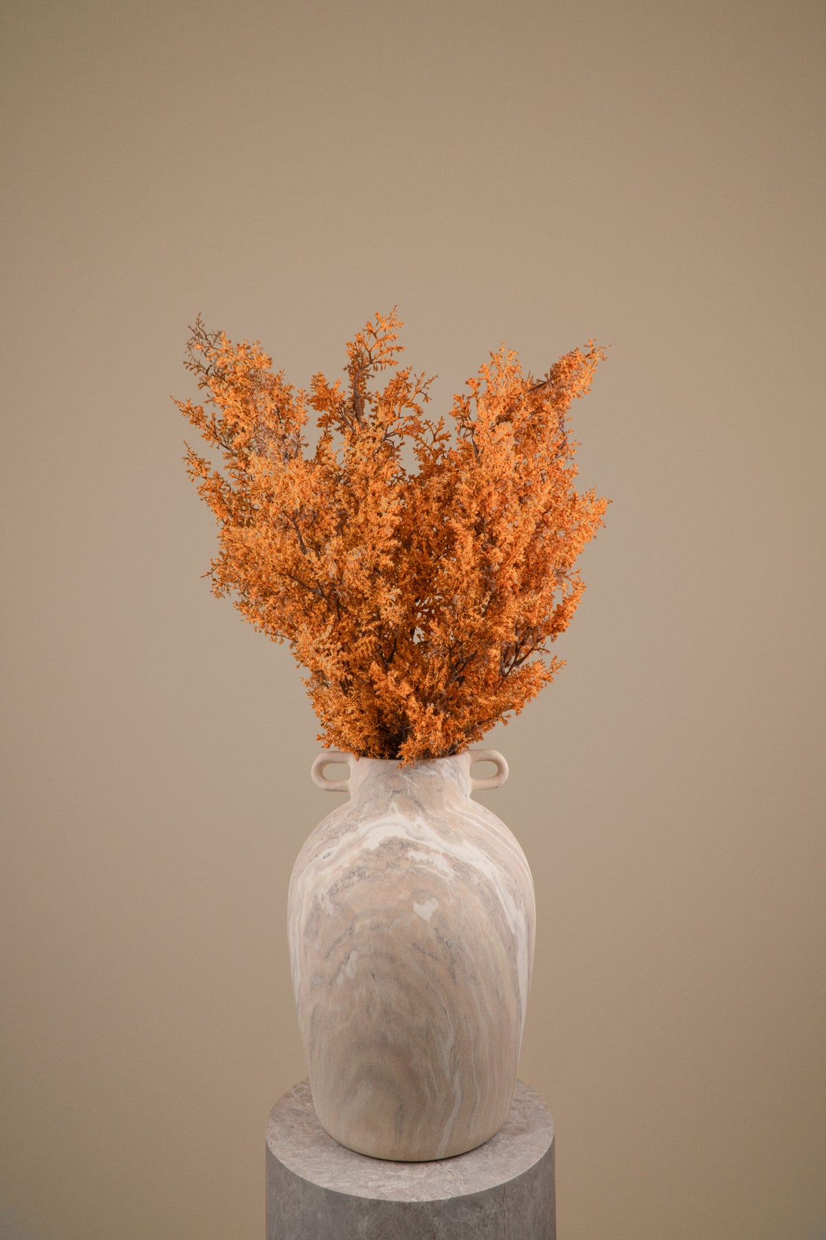 A dried orange botanical arrangement in a light beige marbled ceramic vase with small side handles, displayed on a round pedestal against a warm beige studio background.