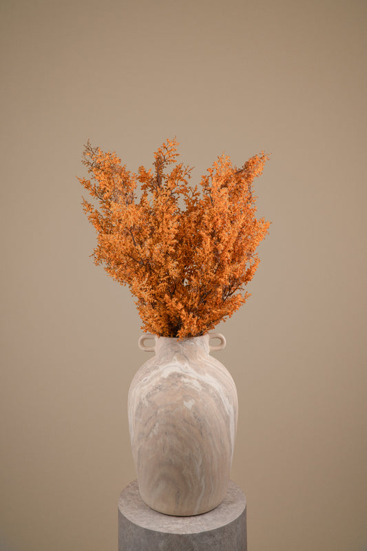 A dried orange botanical arrangement in a light beige marbled ceramic vase with small side handles, displayed on a round pedestal against a warm beige studio background.