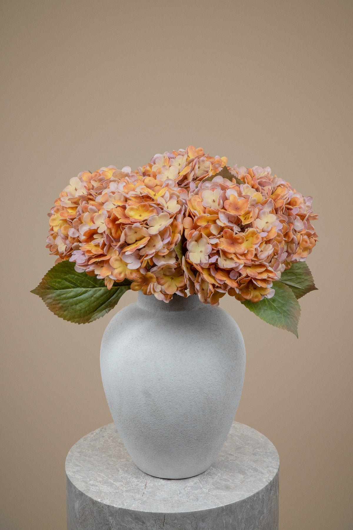 A vase arrangement featuring dried orange and pinkish hydrangea blooms with green leaves, displayed in a matte gray ceramic vase on a round stone pedestal against a solid beige background.
