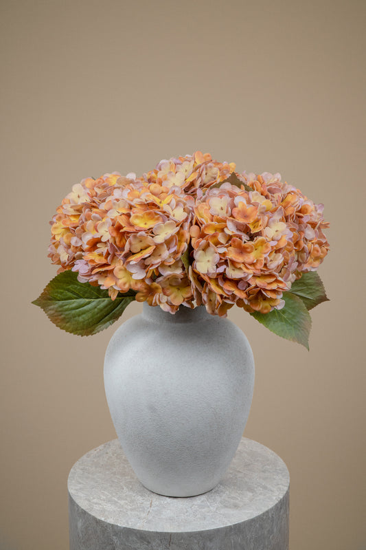A vase arrangement featuring dried orange and pinkish hydrangea blooms with green leaves, displayed in a matte gray ceramic vase on a round stone pedestal against a solid beige background.