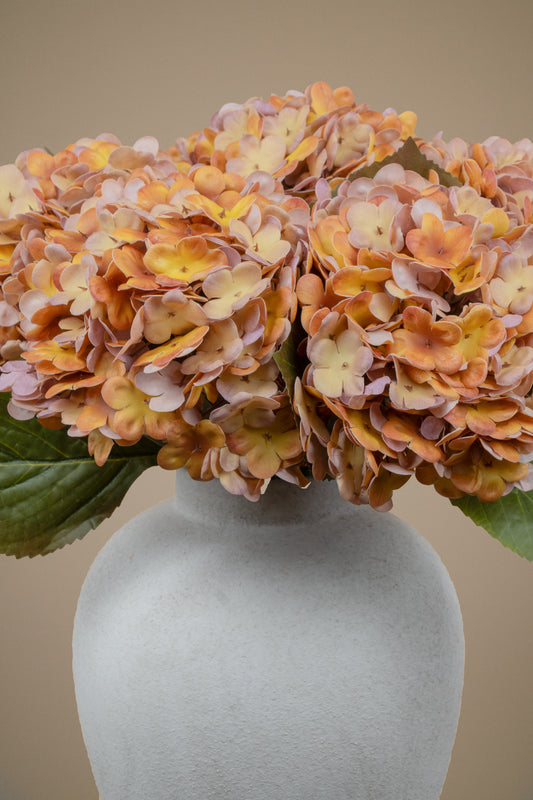 A close-up of a faux hydrangea flower arrangement in a matte light gray vase against a warm beige background.