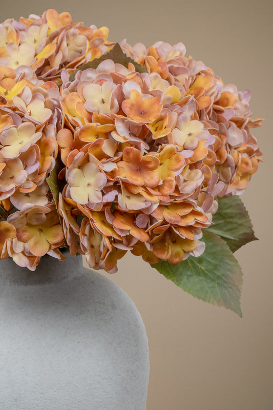 A close-up of a faux hydrangea flower arrangement in a matte gray vase against a solid beige background.