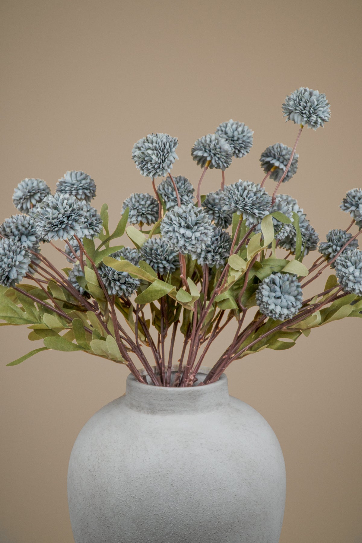 A bouquet of artificial dried flowers with dusty blue-gray pom-pom blooms and green leaves, arranged in a textured light gray ceramic vase against a solid beige background.