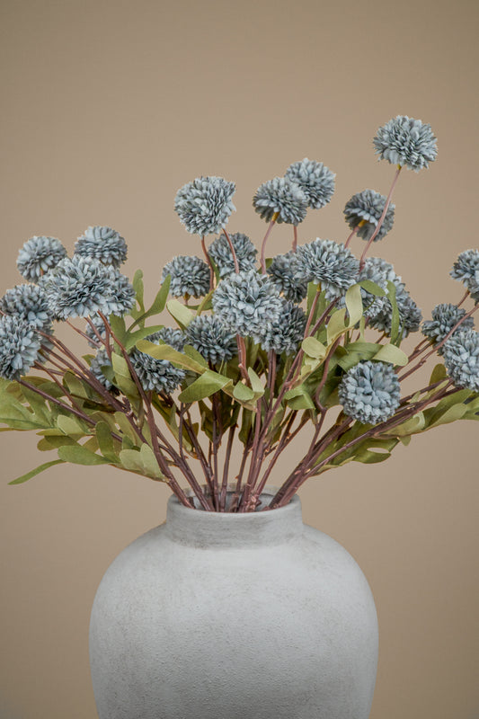 A bouquet of artificial dried flowers with dusty blue-gray pom-pom blooms and green leaves, arranged in a textured light gray ceramic vase against a solid beige background.