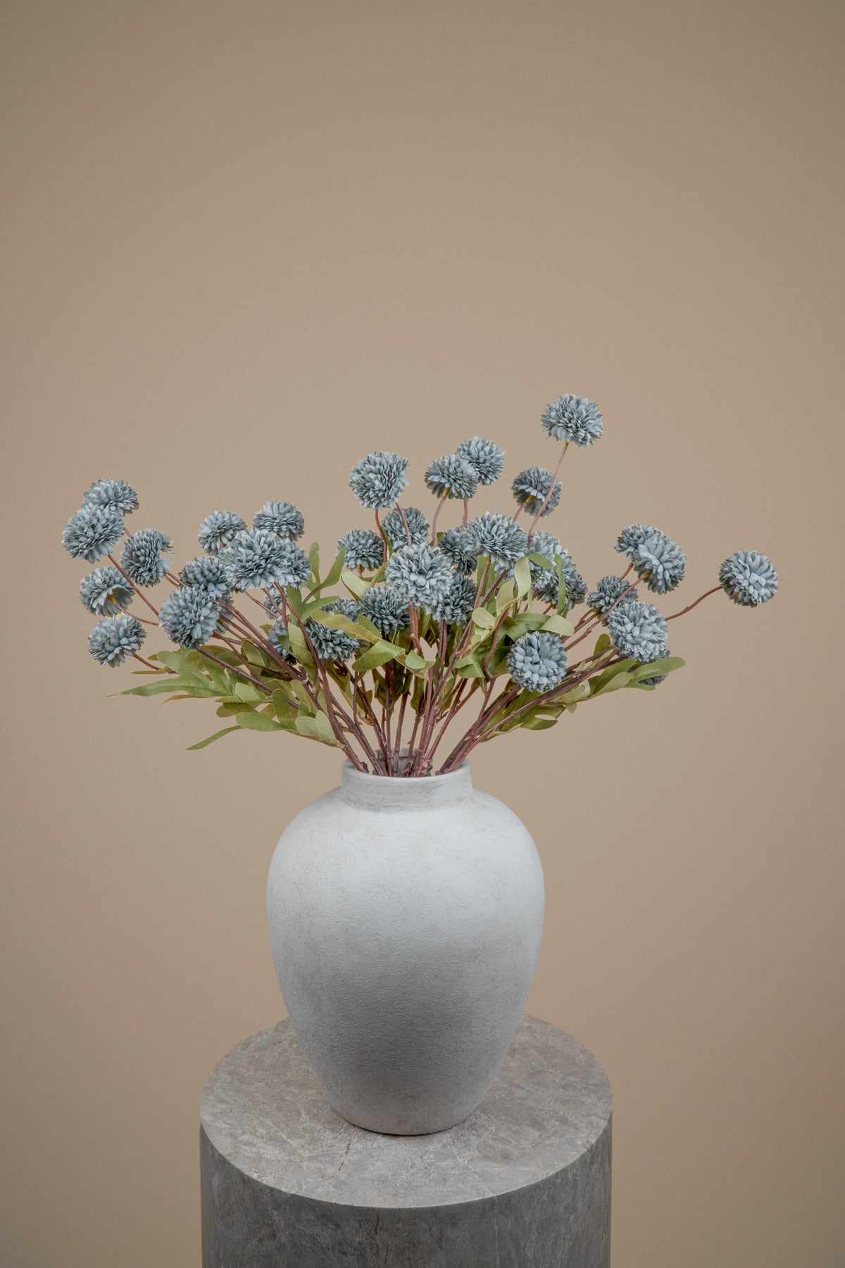 A white ceramic vase filled with dried blue globe flowers and green leaves, displayed on a round gray pedestal against a solid beige background.