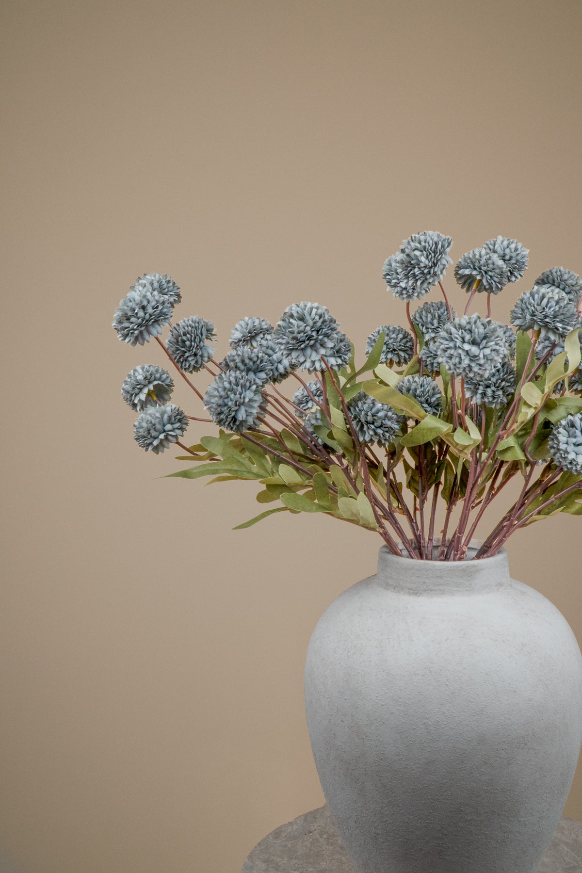 A bouquet of dried blue-gray pom-pom flowers with green leaves arranged in a matte light gray ceramic vase against a solid warm beige background.