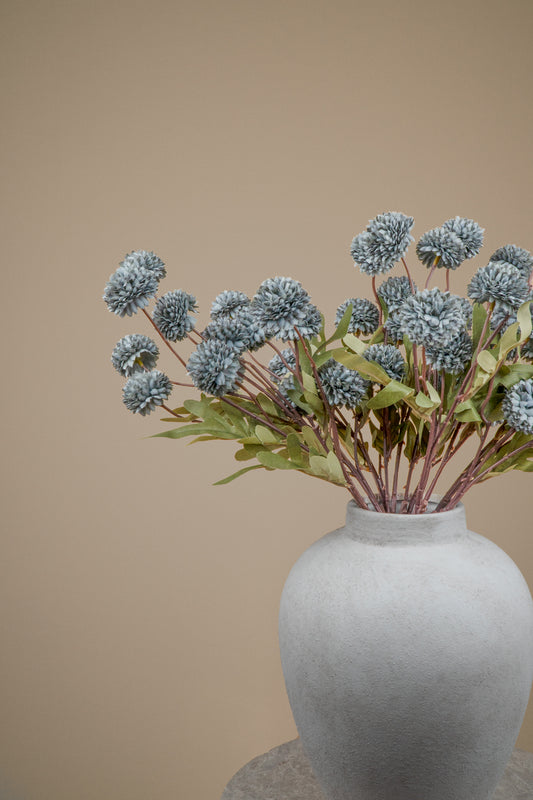 A bouquet of dried blue-gray pom-pom flowers with green leaves arranged in a matte light gray ceramic vase against a solid warm beige background.