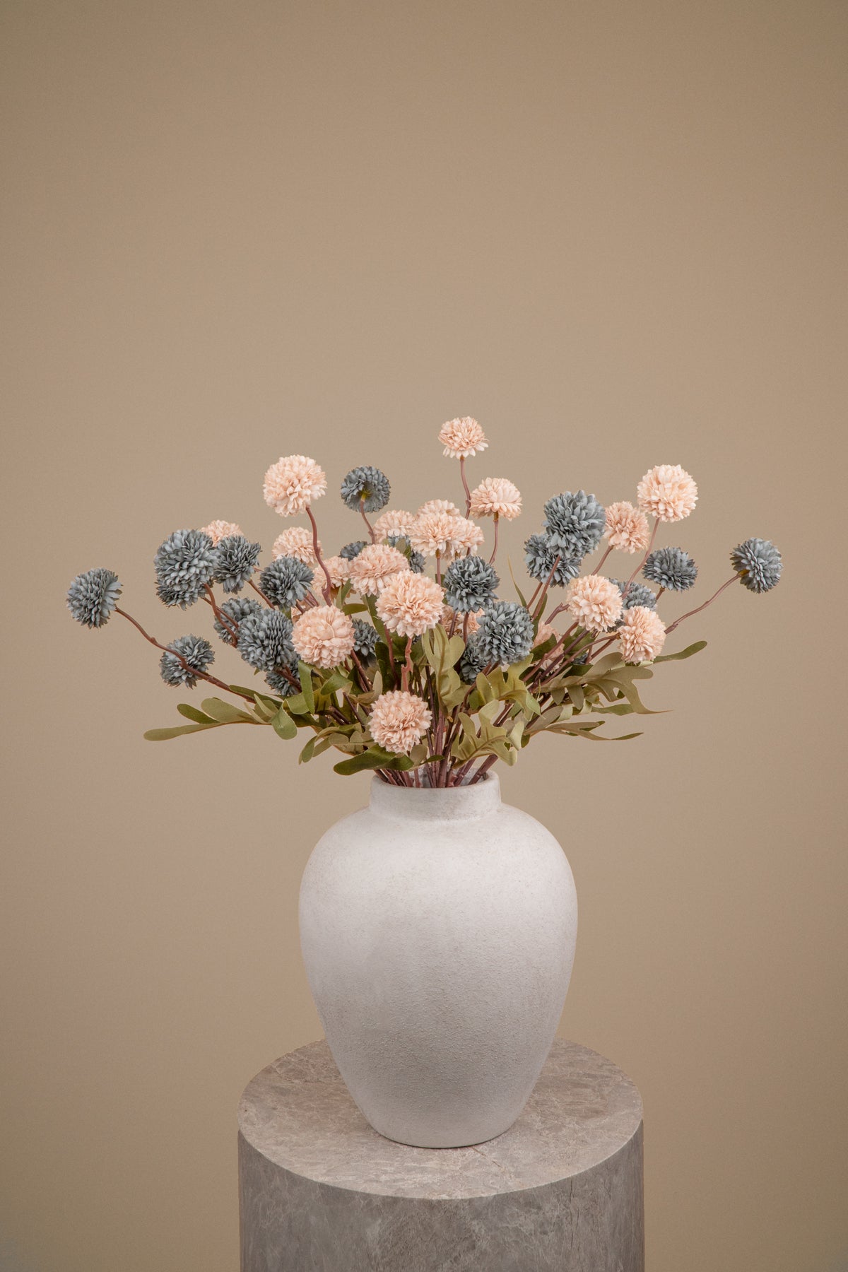 An artificial floral arrangement featuring dusty pink and slate blue pom-pom flowers with green foliage, presented in a matte white ceramic vase on a round stone pedestal against a solid beige background.