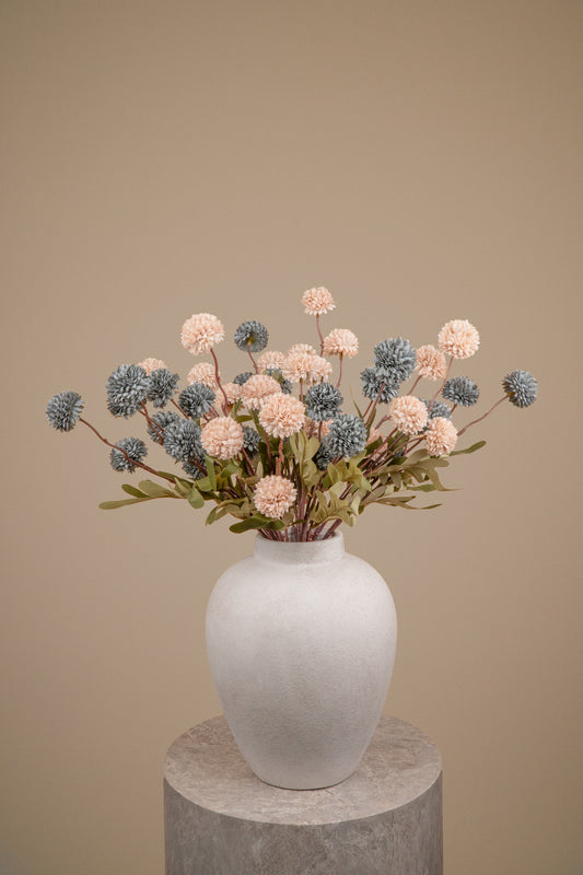 An artificial floral arrangement featuring dusty pink and slate blue pom-pom flowers with green foliage, presented in a matte white ceramic vase on a round stone pedestal against a solid beige background.