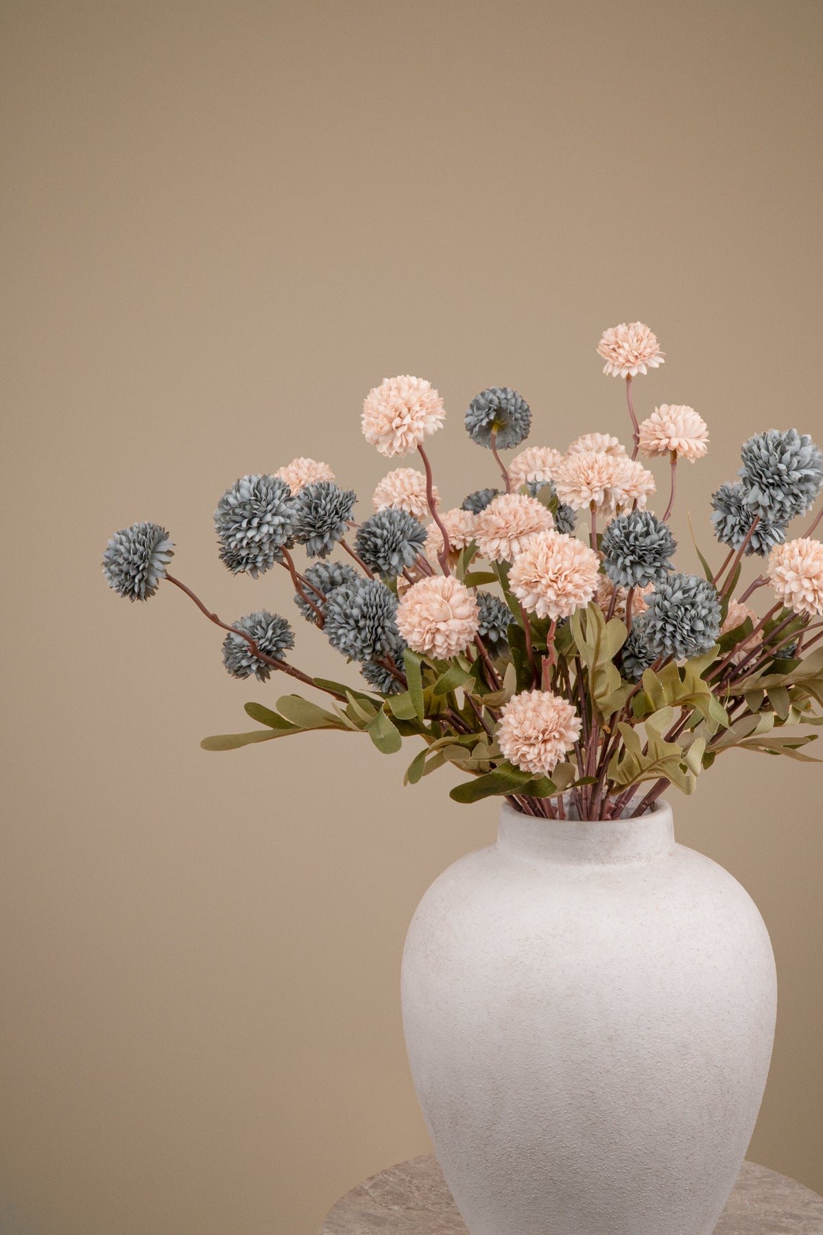 A dried floral arrangement featuring dusty pink and slate blue pom-pom flowers with green leaves, presented in a textured off-white ceramic vase against a solid beige background.