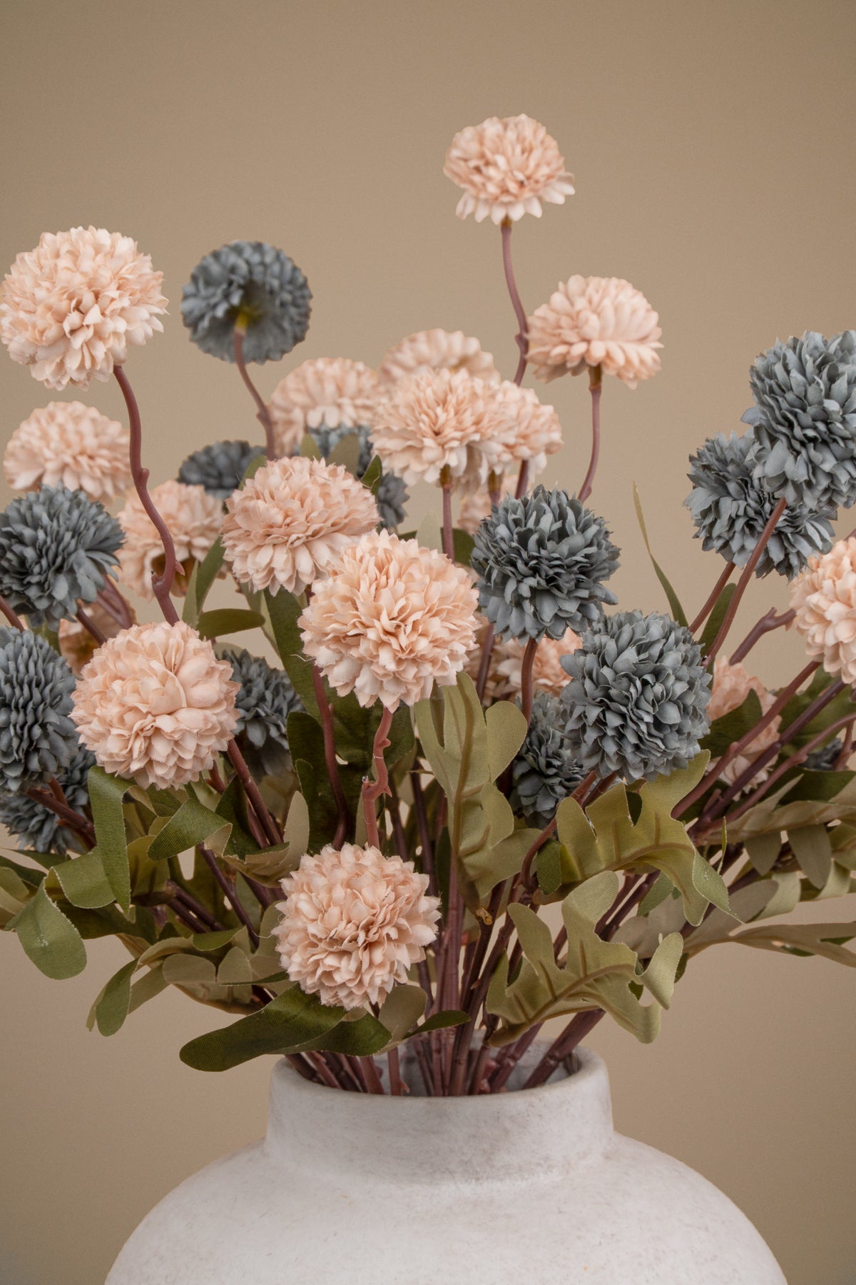 A bouquet of artificial dried flowers in soft blush pink, muted blue-gray, and olive green tones, arranged in a textured off-white ceramic vase against a warm beige background.