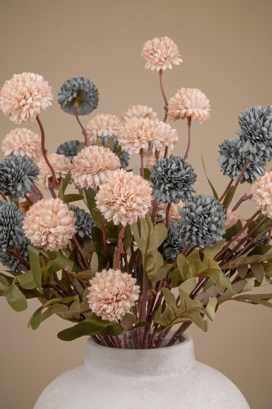 A bouquet of artificial dried flowers in soft blush pink, muted blue-gray, and olive green tones, arranged in a textured off-white ceramic vase against a warm beige background.