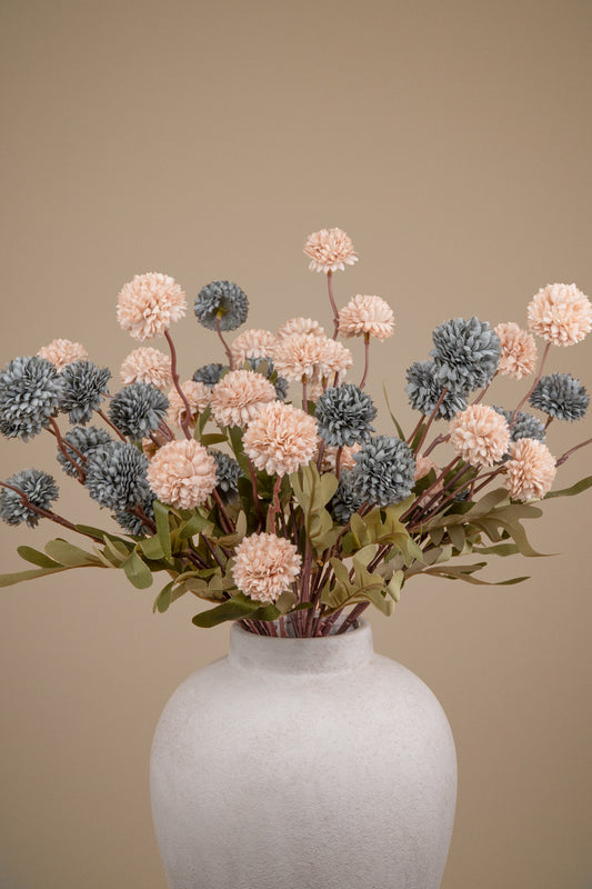 An artificial floral arrangement featuring dusty blush pink and slate blue pom-pom flowers with green leaves, presented in a matte white ceramic vase against a solid beige background.
