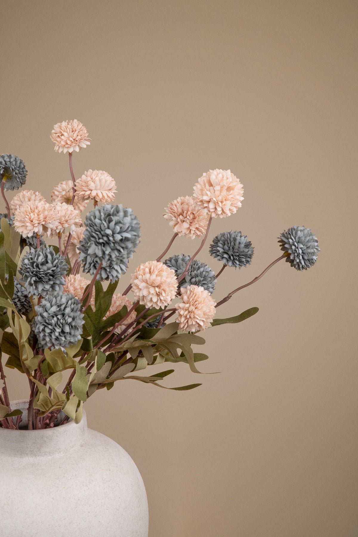 A dried floral arrangement featuring textured pom-pom flowers in blush pink and slate blue-gray tones, with muted green leaves, presented in a matte white ceramic vase against a solid warm beige background.