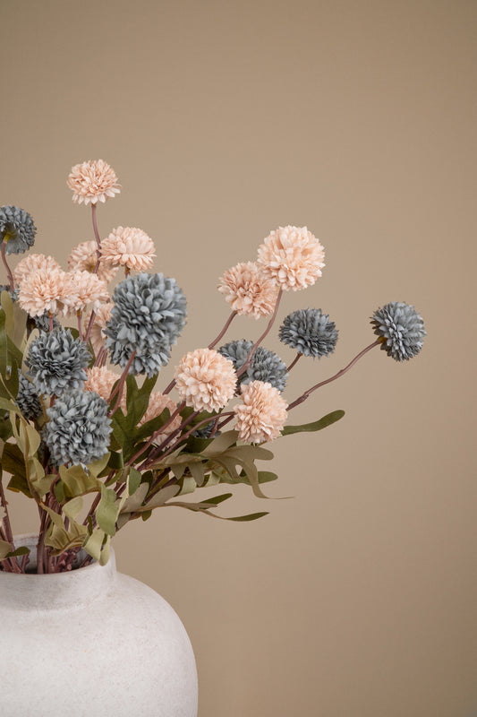 A dried floral arrangement featuring textured pom-pom flowers in blush pink and slate blue-gray tones, with muted green leaves, presented in a matte white ceramic vase against a solid warm beige background.