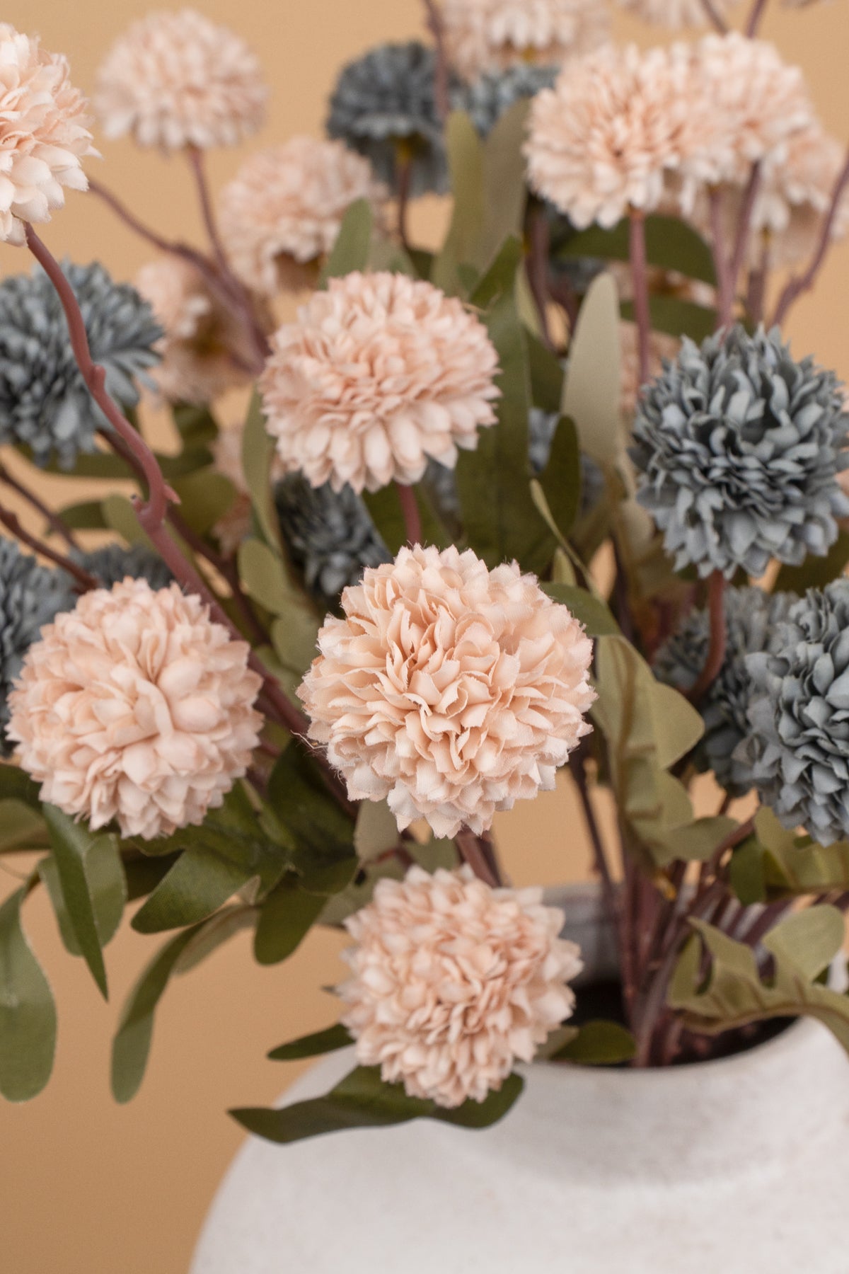 A close-up of a bouquet of artificial flowers in a white ceramic vase, featuring ruffled blush peach blooms, muted blue-gray flowers, and green leaves against a warm beige background.