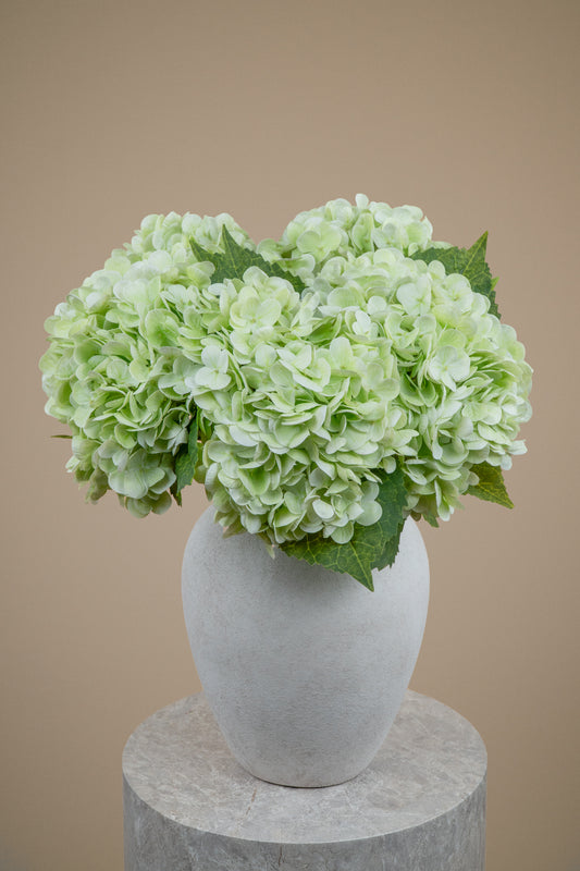 Light green hydrangea flower arrangement in a matte white ceramic vase, displayed on a round stone pedestal against a solid beige background.