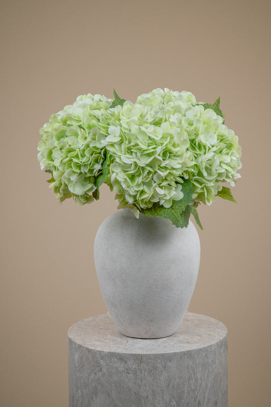 Light green hydrangea flowers arranged in a matte white ceramic vase, displayed on a round gray pedestal against a solid beige background.