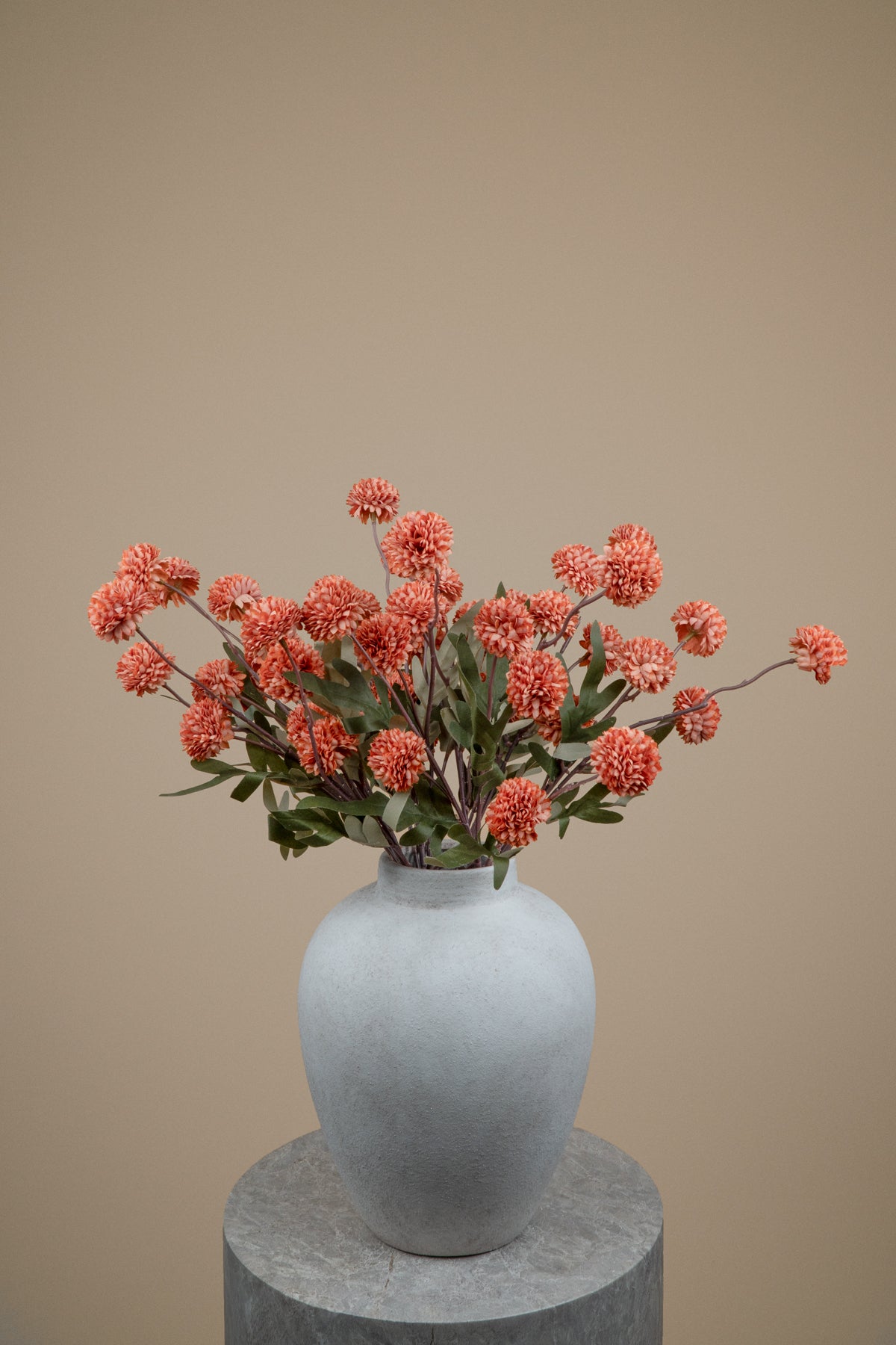 A vase arrangement of coral-pink dried flowers with green leaves in a matte light gray ceramic vase, displayed on a round gray pedestal against a solid beige background.