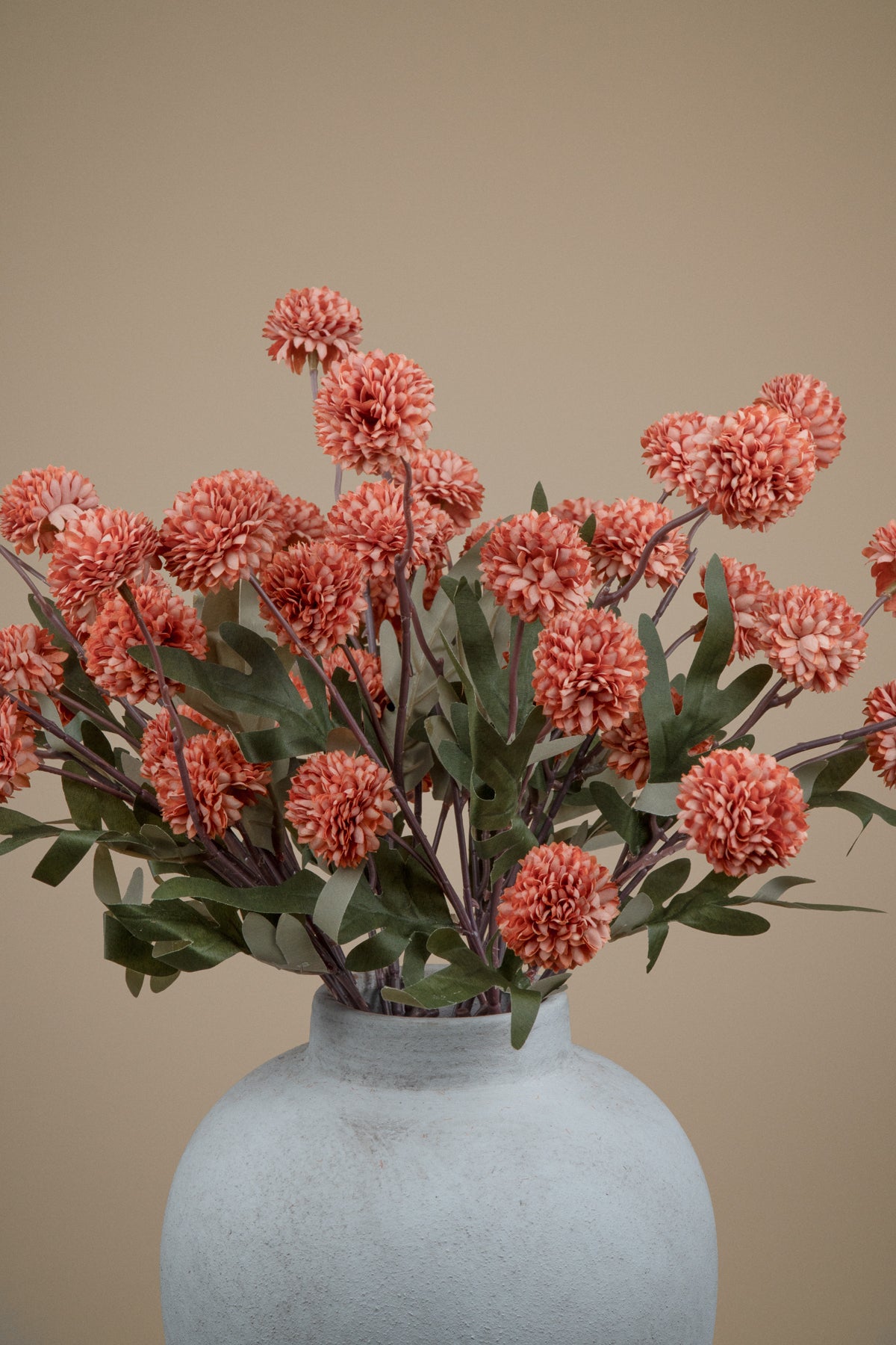 A bouquet of coral-pink chrysanthemum-like flowers with green leaves arranged in a matte light gray ceramic vase against a plain warm beige background.