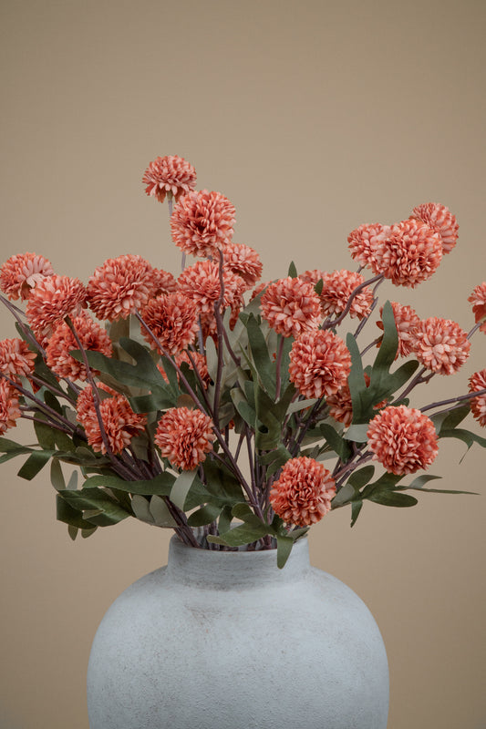 A bouquet of coral-pink chrysanthemum-like flowers with green leaves arranged in a matte light gray ceramic vase against a plain warm beige background.