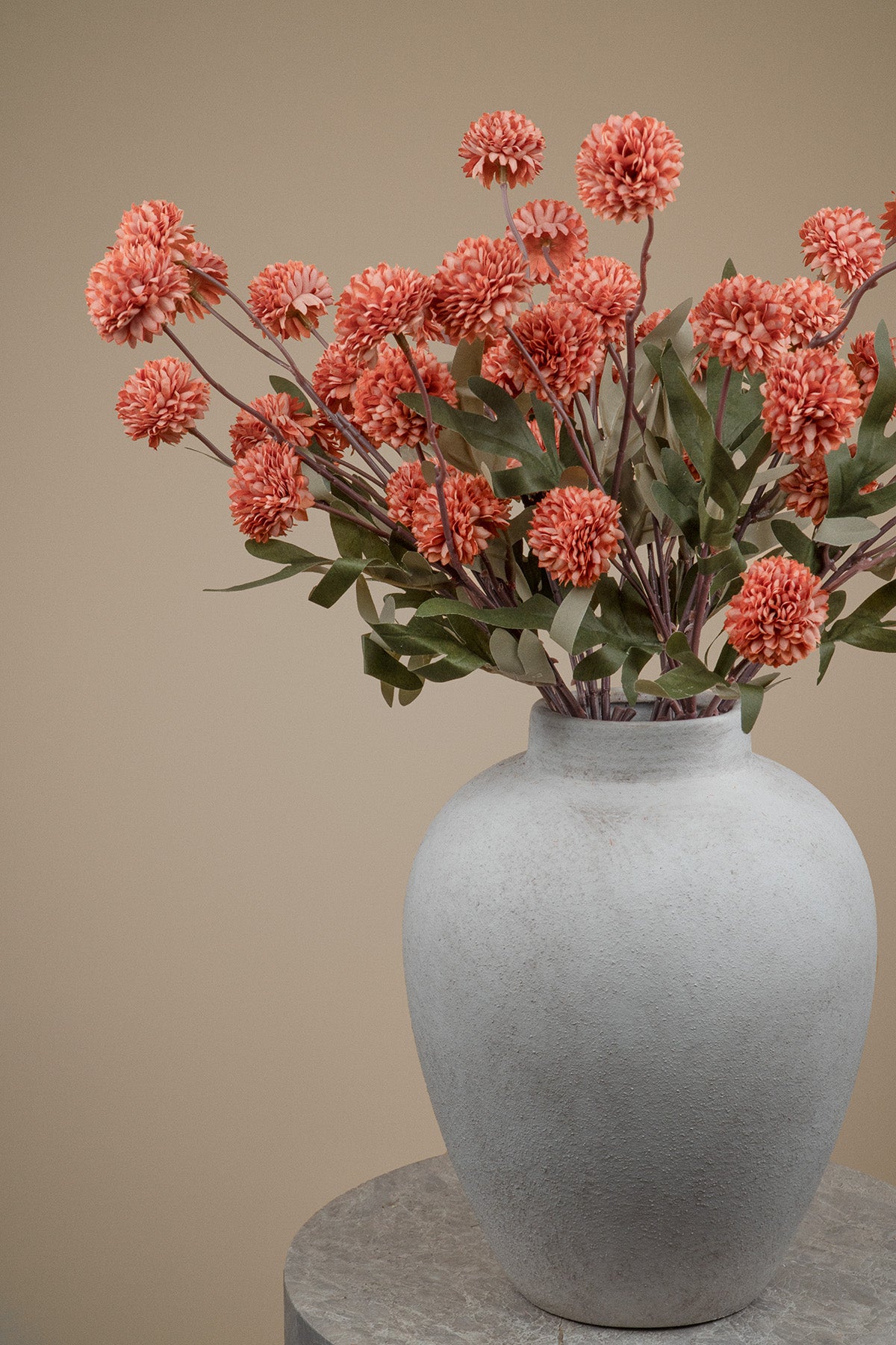 A textured light gray ceramic vase filled with coral-pink chrysanthemum-like flowers and green leaves, set against a warm beige background.