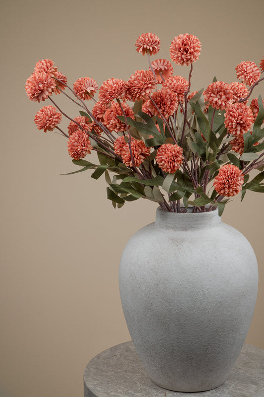 A textured light gray ceramic vase filled with coral-pink chrysanthemum-like flowers and green leaves, set against a warm beige background.
