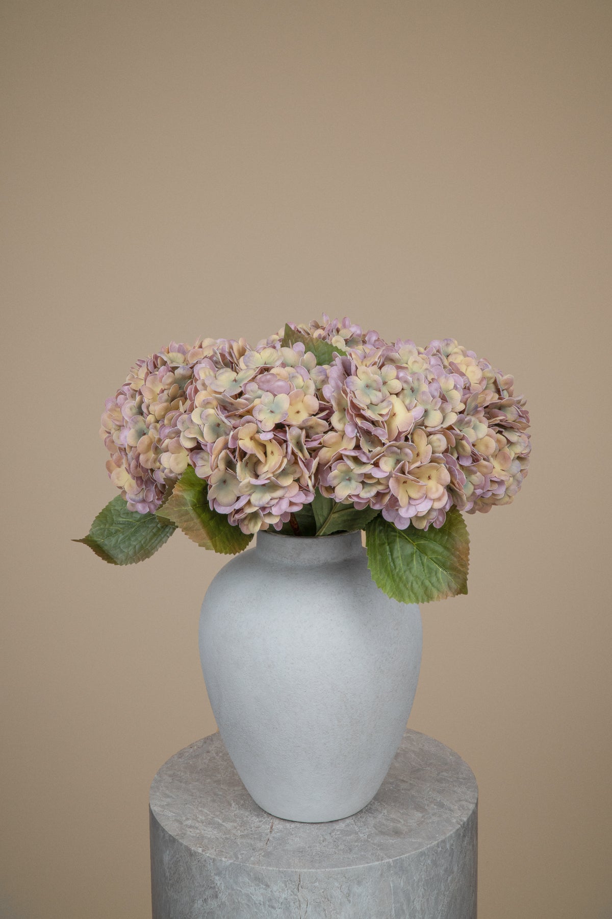 A bouquet of dried or artificial hydrangea flowers in muted pink, beige, and green tones, arranged in a matte light gray ceramic vase on a round stone pedestal against a plain beige background.