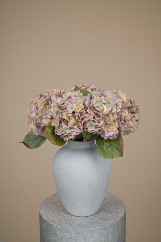 A bouquet of dried or artificial hydrangea flowers in muted pink, beige, and green tones, arranged in a matte light gray ceramic vase on a round stone pedestal against a plain beige background.