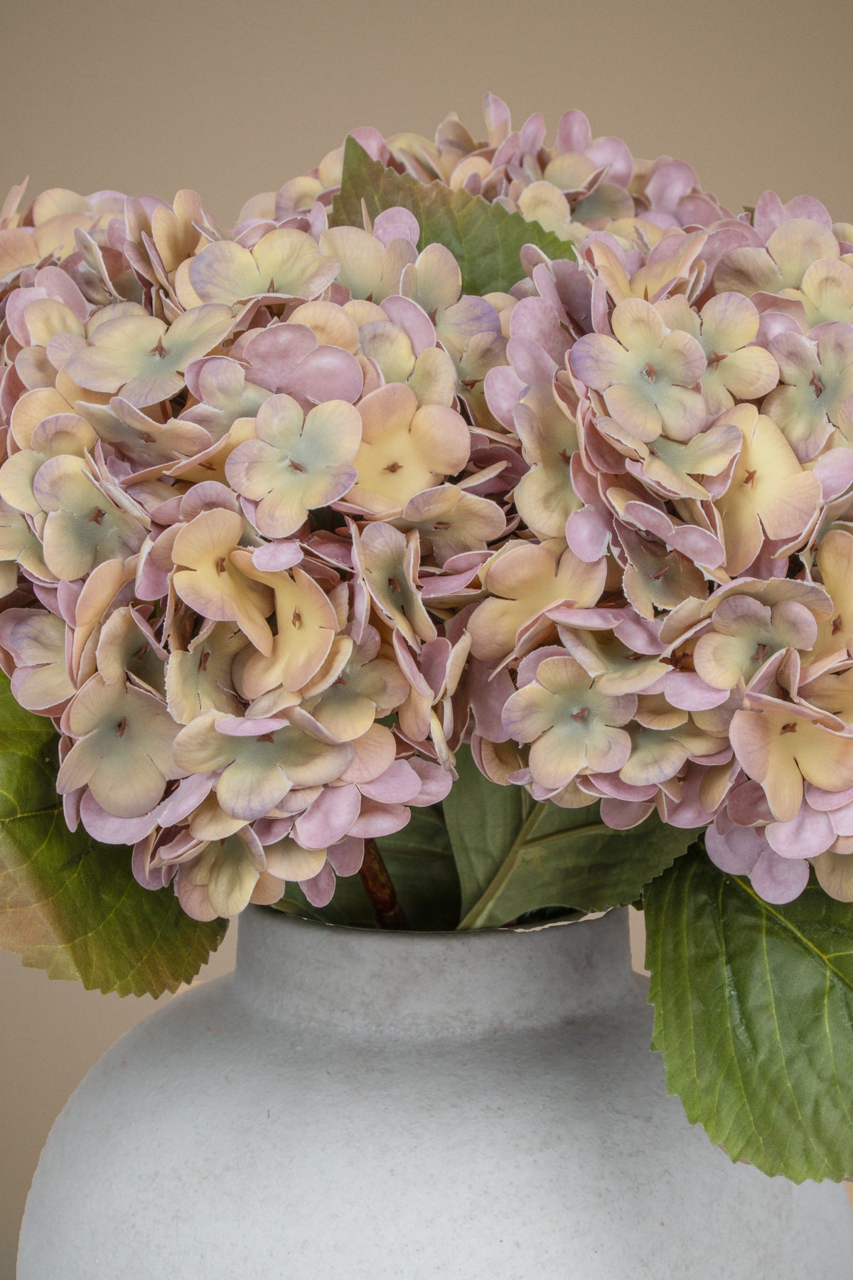 A close-up of a faux hydrangea arrangement in a matte white ceramic vase against a solid beige background.