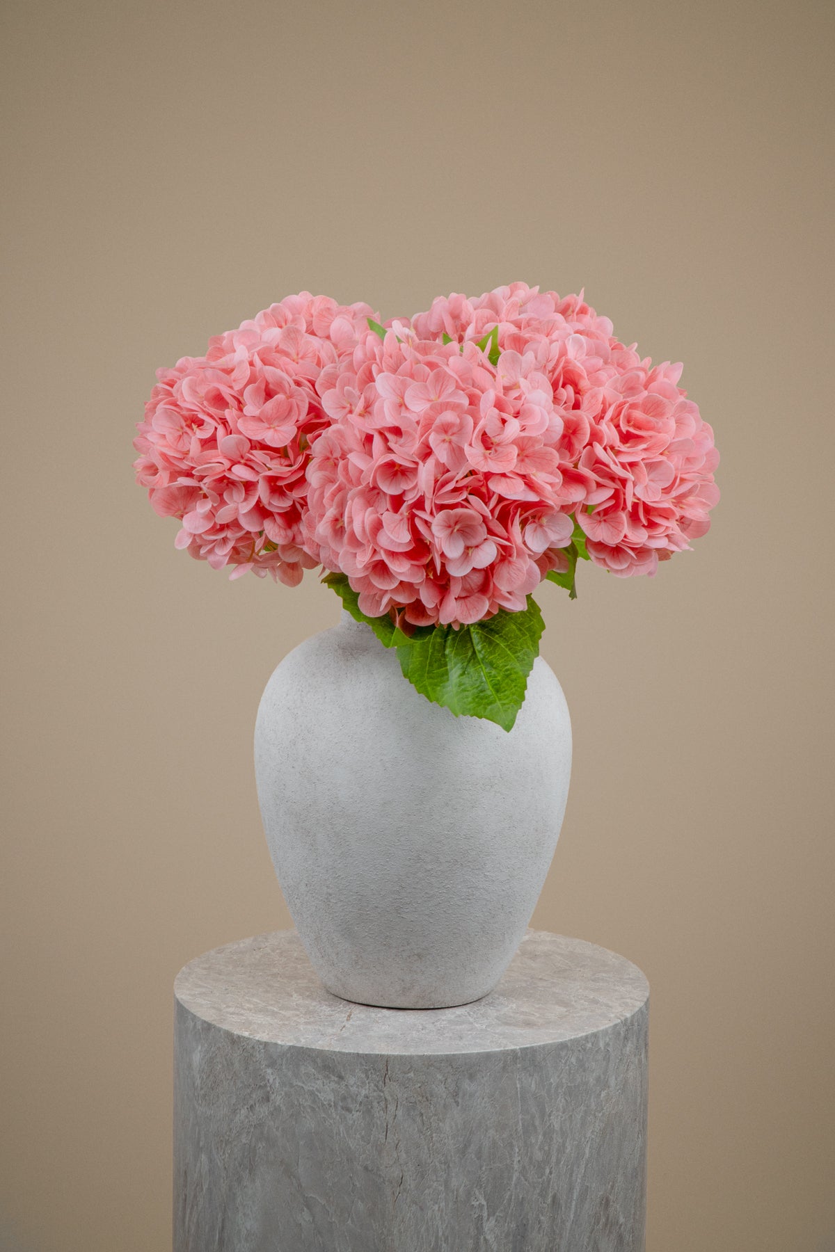 Pink artificial hydrangea flowers arranged in a white ceramic vase, displayed on a gray stone pedestal against a solid beige background.