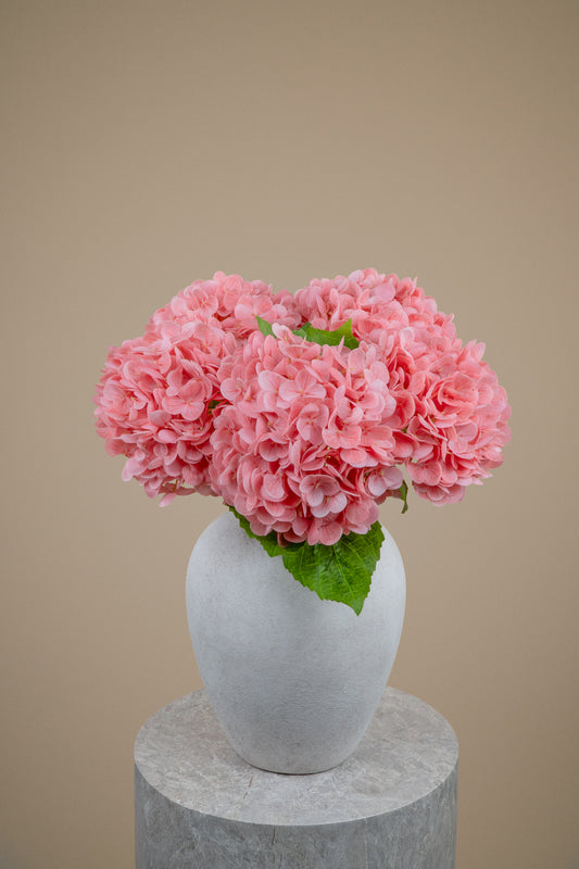 Three pink hydrangea flower bouquets with green leaves arranged in a white ceramic vase, set on a round gray stone pedestal against a solid beige background.
