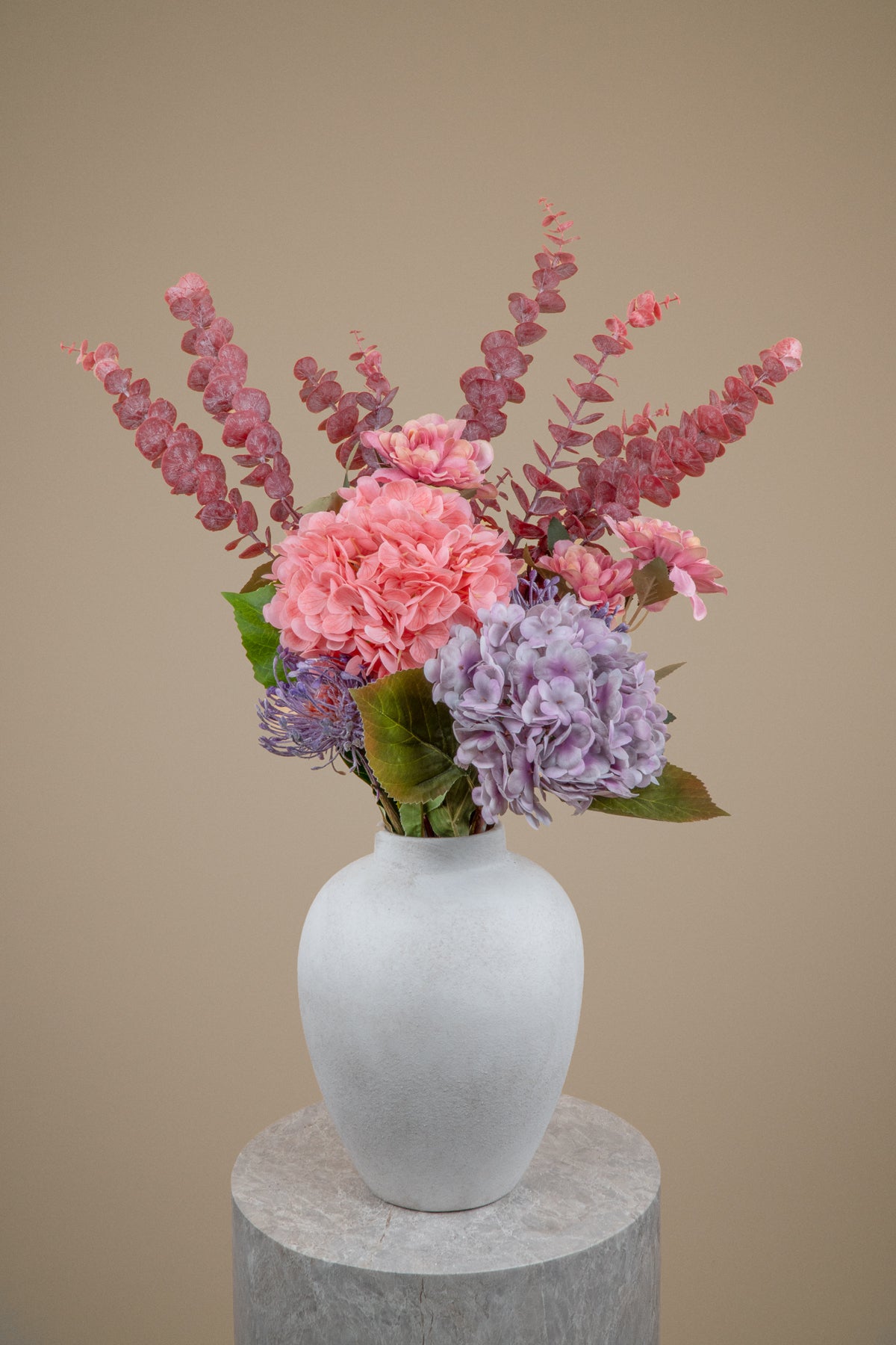 A faux flower bouquet with pink and purple hydrangeas and dusty rose eucalyptus stems arranged in a matte white ceramic vase, set on a round gray pedestal against a solid beige background.