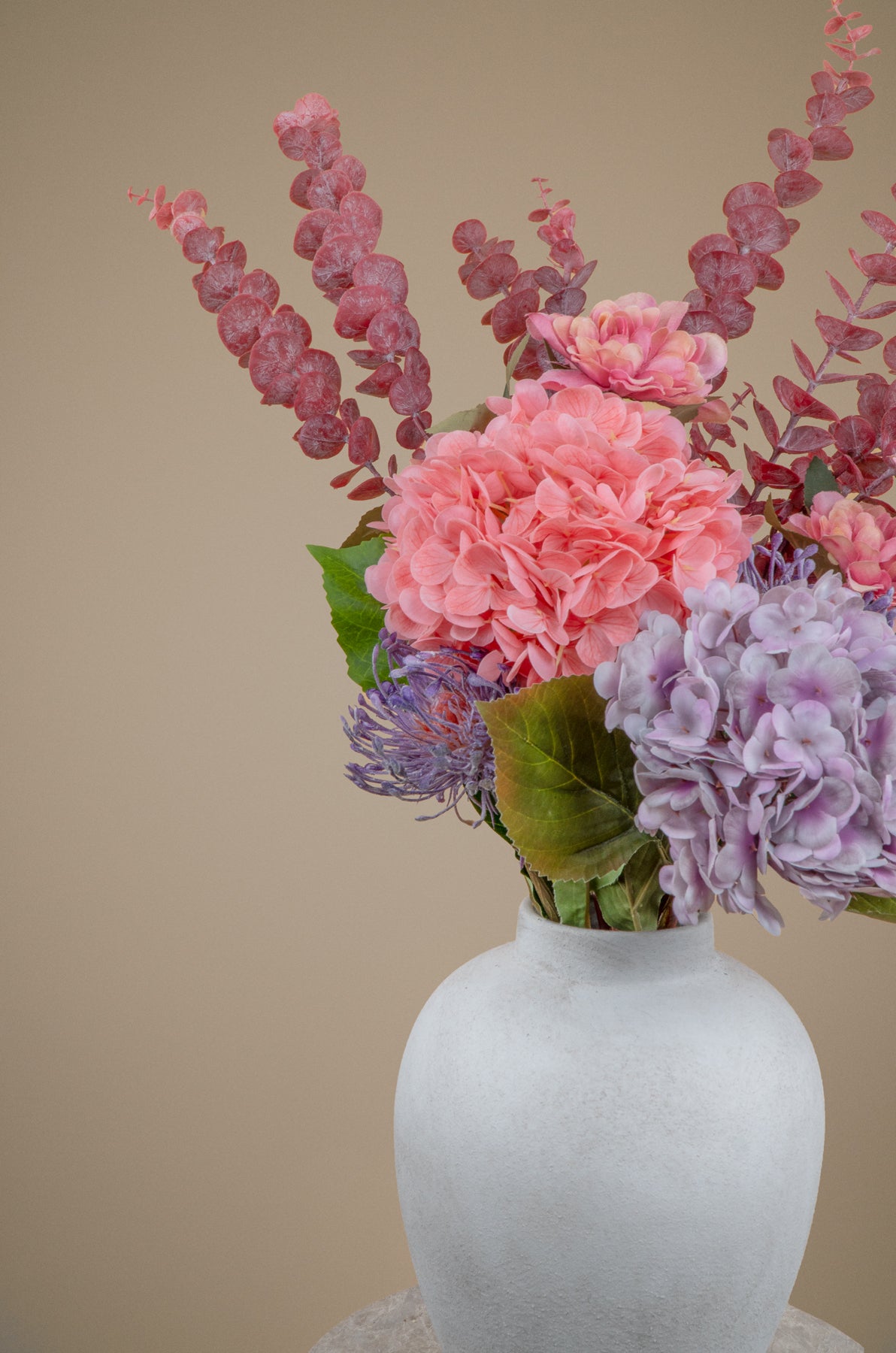 A floral arrangement featuring pink and purple hydrangeas, dusty rose eucalyptus stems, and green leaves, presented in a textured white ceramic vase against a solid beige background.