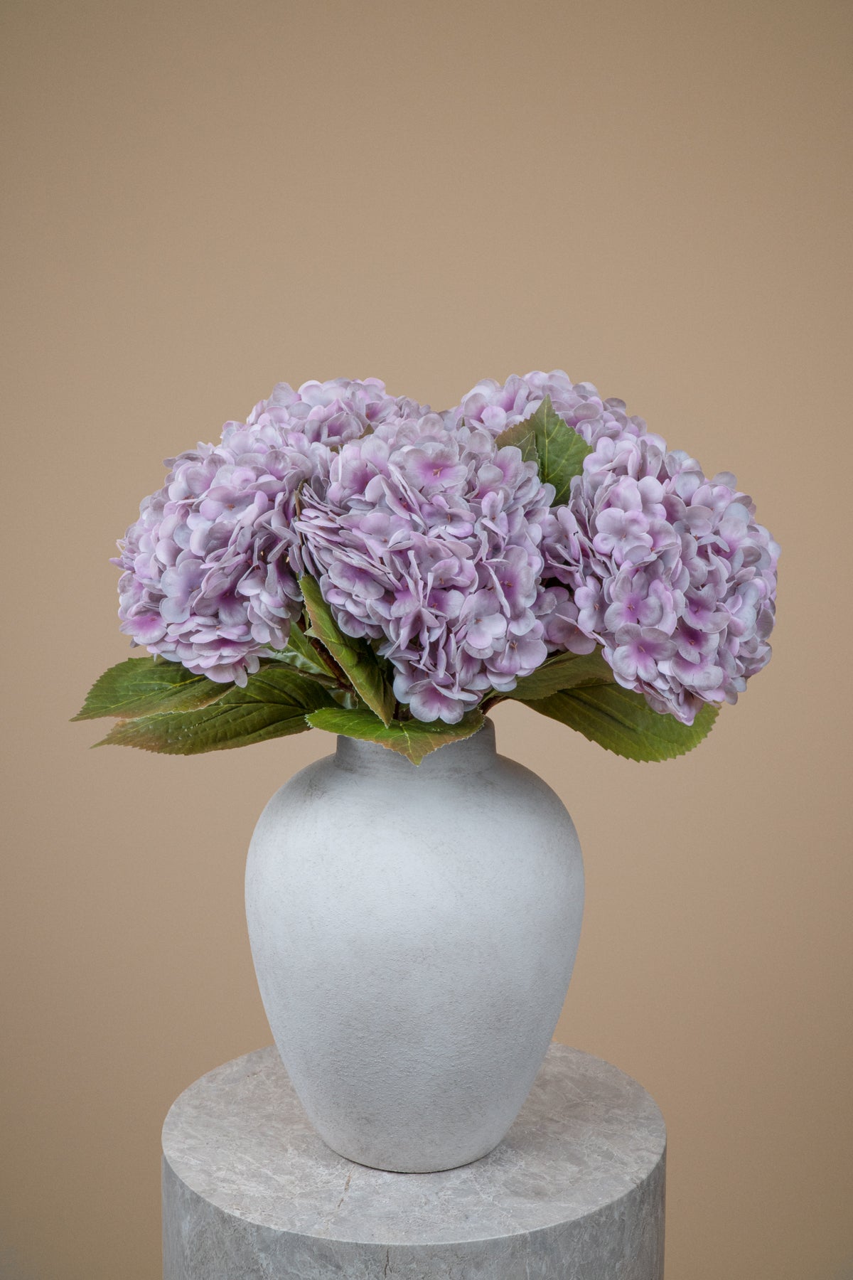 A bouquet of light purple hydrangea flowers with green leaves arranged in a matte white ceramic vase, set on a round stone pedestal against a solid beige background.