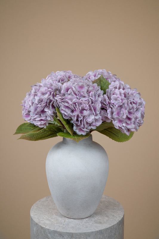 A bouquet of light purple hydrangea flowers with green leaves arranged in a matte white ceramic vase, set on a round stone pedestal against a solid beige background.