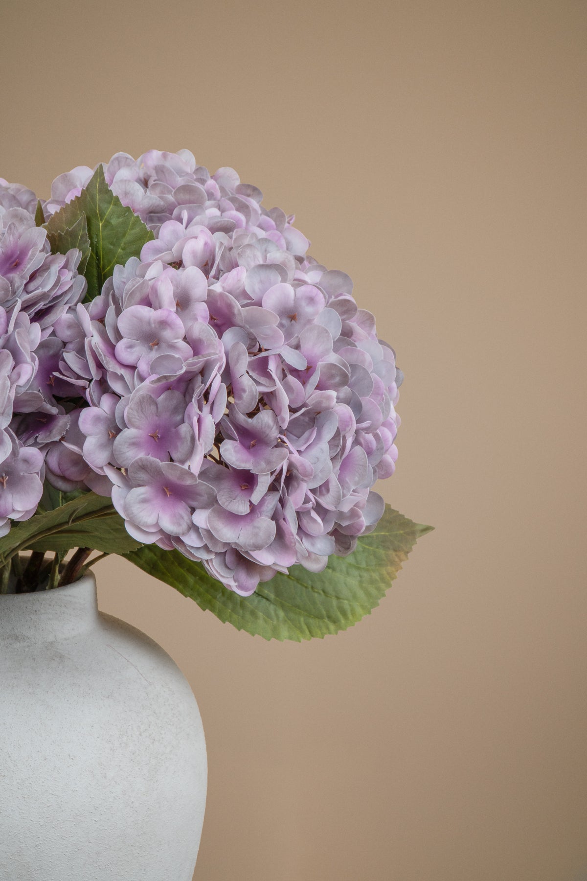 A close-up of a pale lavender-purple hydrangea bouquet in a textured off-white ceramic vase against a solid warm beige background.