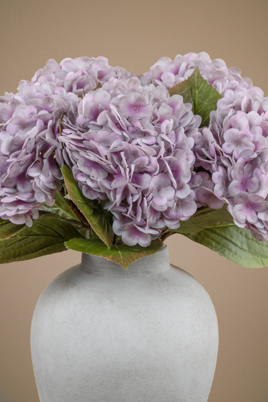 A bouquet of light purple hydrangea flowers with green leaves arranged in a matte white ceramic vase against a solid beige background.