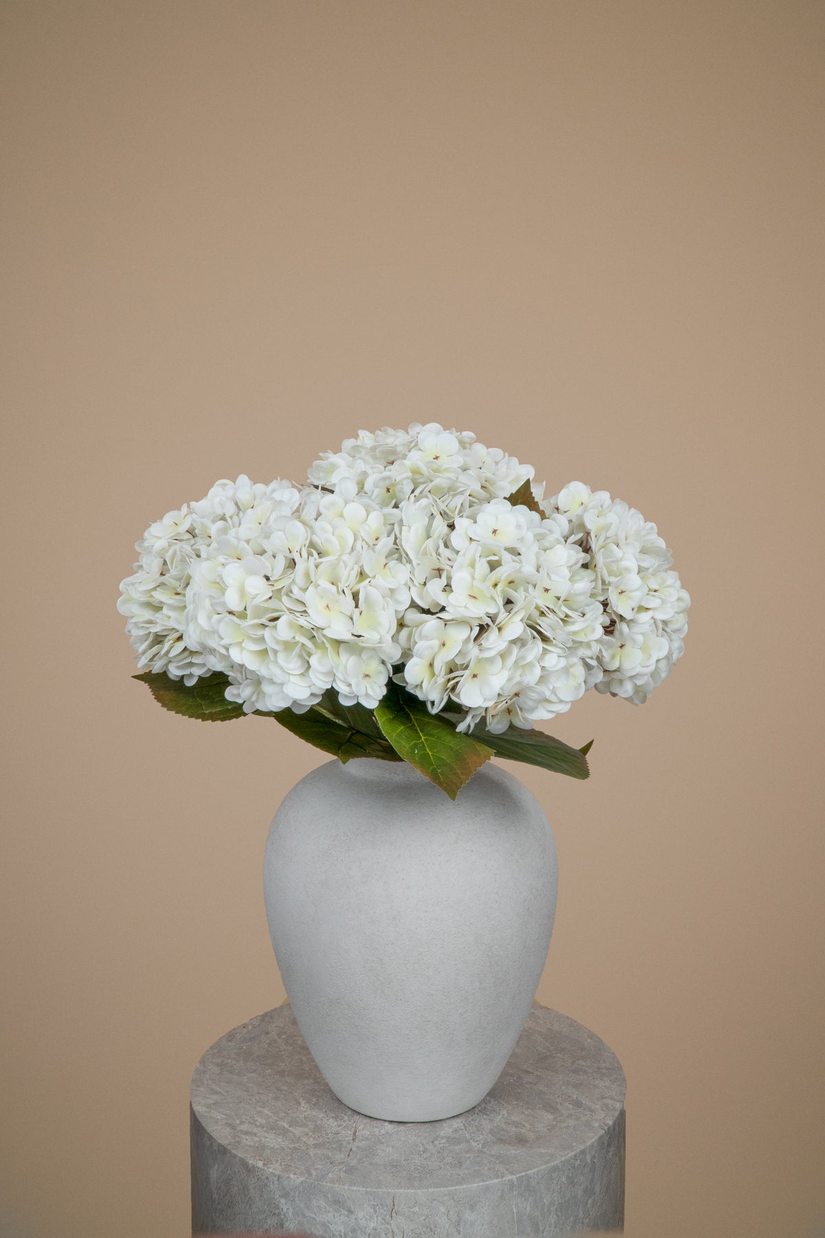 White hydrangea flowers arranged in a matte light gray ceramic vase, set on a round gray pedestal against a solid warm beige background.