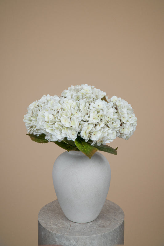 White hydrangea flowers arranged in a matte light gray ceramic vase, set on a round gray pedestal against a solid warm beige background.