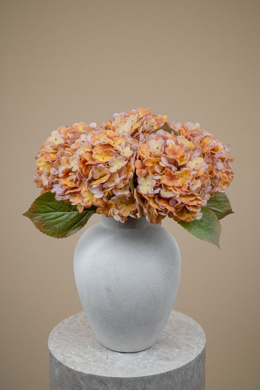 A vase arrangement featuring dried orange and pinkish hydrangea blooms with green leaves, displayed in a matte gray ceramic vase on a round stone pedestal against a solid beige background.
