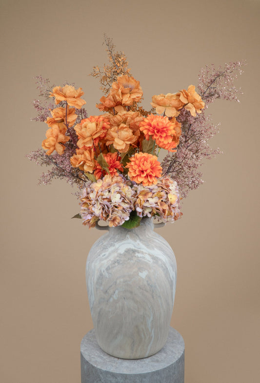 An artificial floral arrangement featuring orange and peach roses, dahlia-like blooms, and light purple filler stems, displayed in a gray marble-patterned ceramic vase on a matching gray pedestal against a solid beige background.