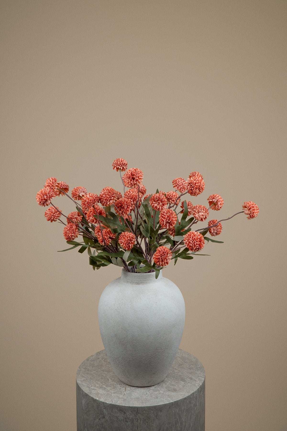 A white ceramic vase filled with orange pom-pom flowers and green leaves, displayed on a round gray pedestal against a solid beige background.