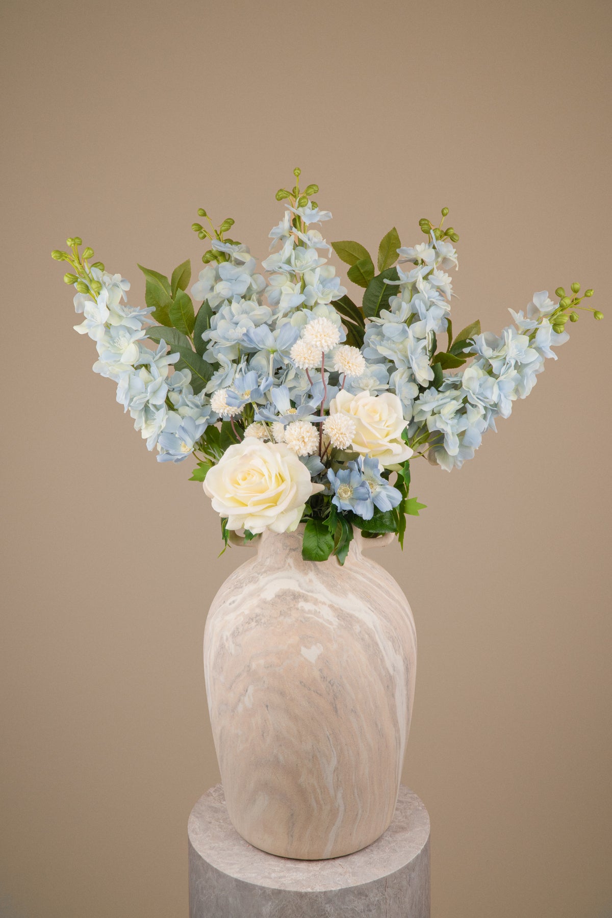 An artificial floral arrangement featuring light blue hydrangeas, white roses, and green foliage, presented in a textured beige and white marbled ceramic vase against a solid beige background.