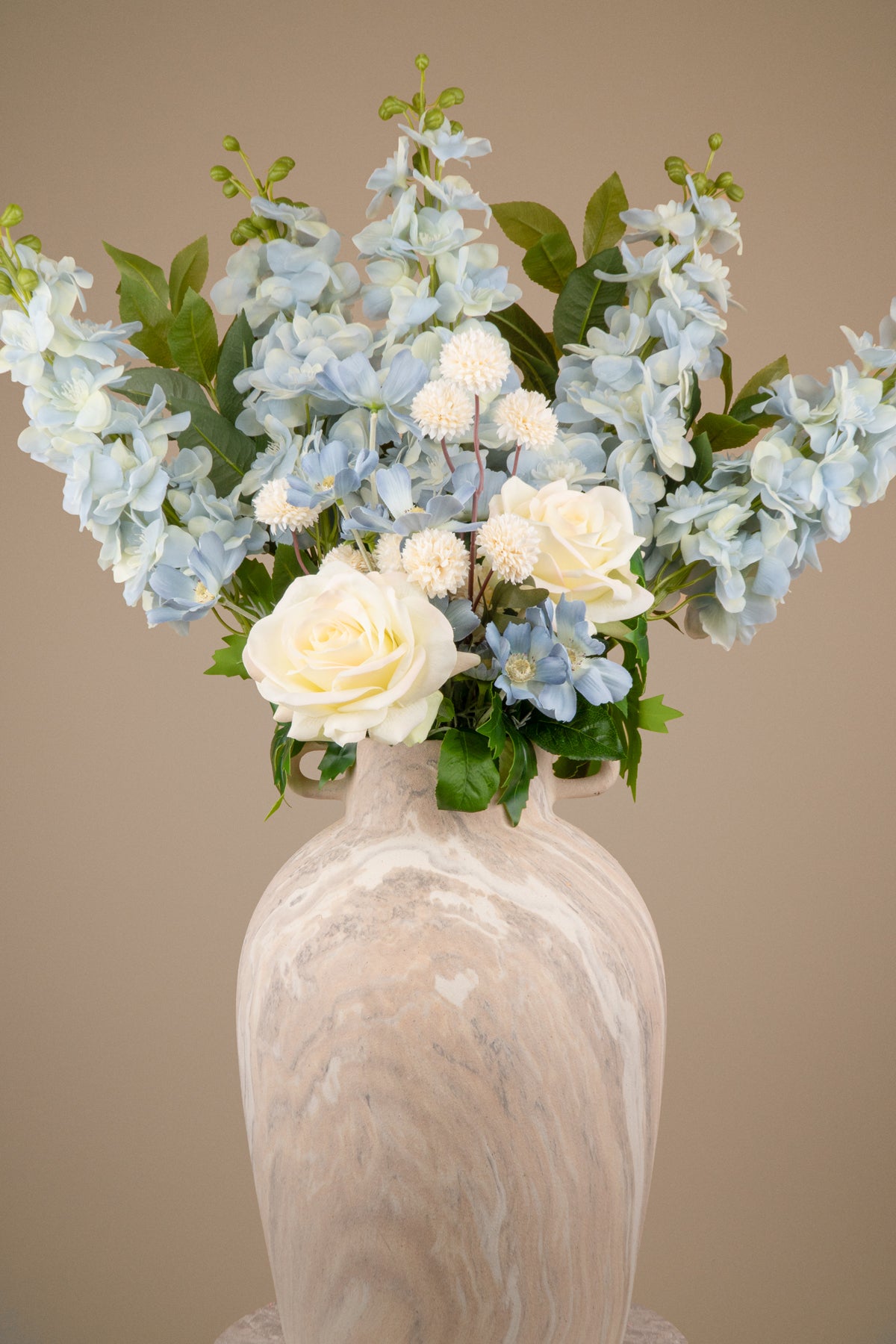 An artificial floral arrangement featuring light blue hydrangeas, white roses, and green foliage, presented in a textured beige and white marbled ceramic vase against a solid beige background.